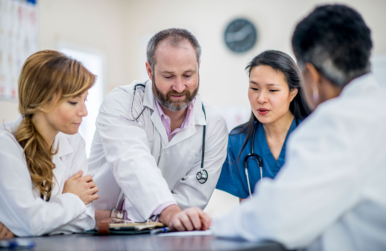 A multi-ethnic group of doctors are indoors in an office. They are gathered around a table to have a meeting. One doctor is pointing to pictures on the table.